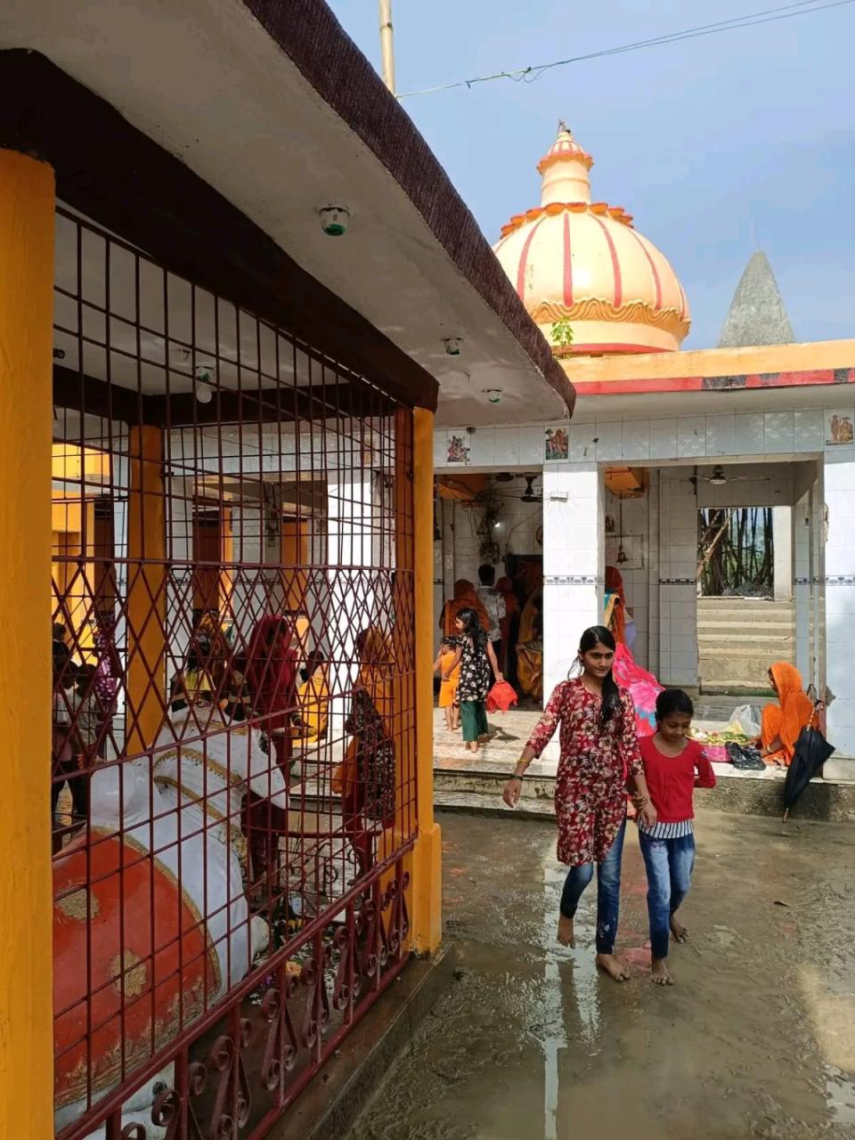 Women leaving the shrine across the wet courtyard of Manara Mahadev Mandir on a monsoon morning, below the striped orange dome