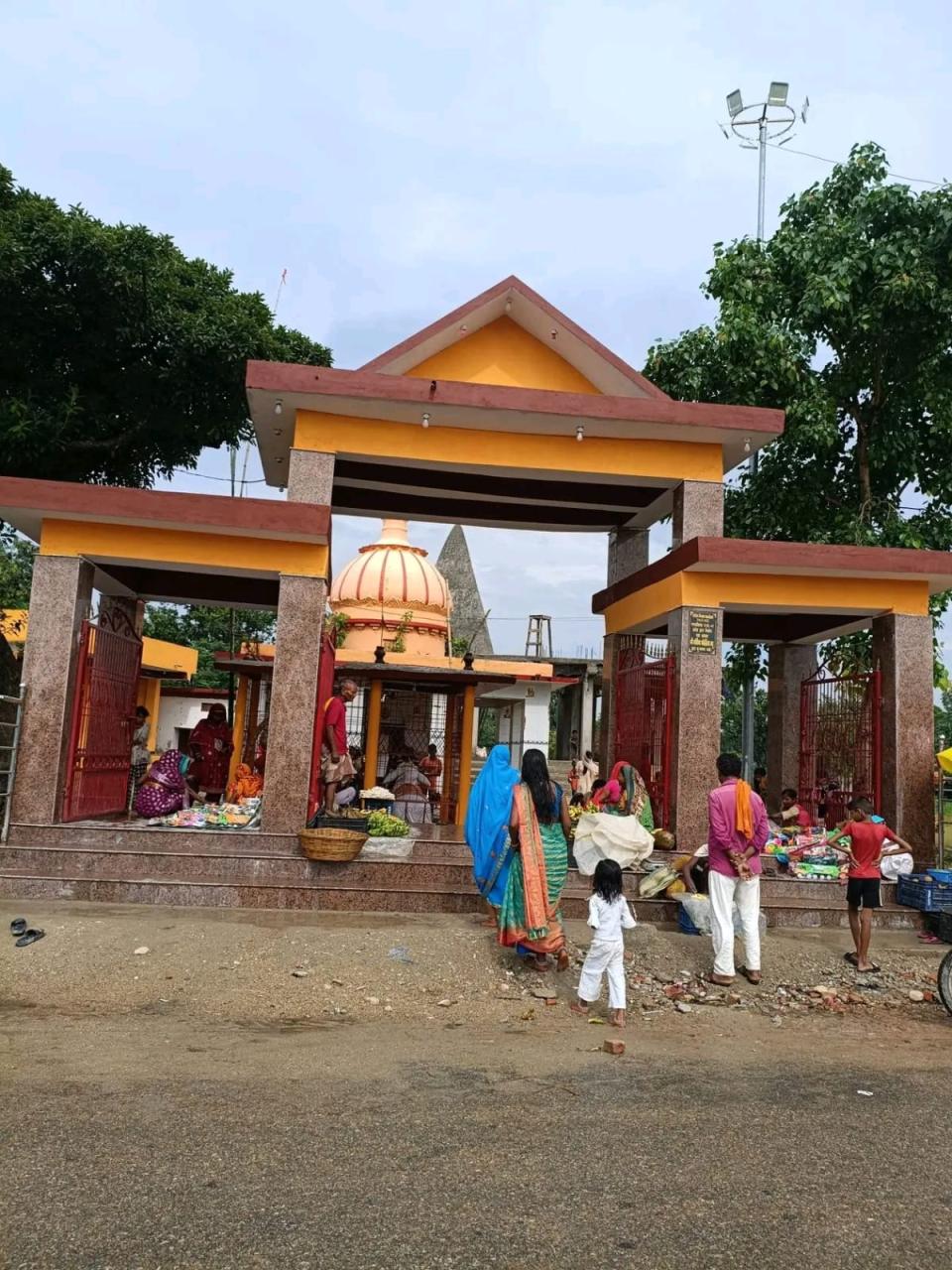 Devotees walking in through the new gateway of Shri Dineshwarnath Mahadev Mandir in Manara, with fruit and puja sellers on the steps and the domed shrine visible through the arch