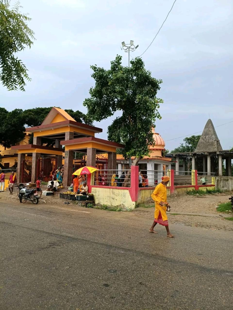 Shri Dineshwarnath Mahadev Mandir seen from the road at Manara, with a devotee in saffron carrying a brass water pot past the fruit stalls